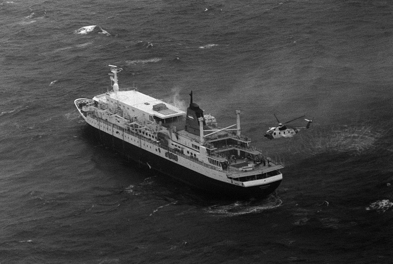 Schwarz-Weiß-Fotografie des in Brand stehenden Kreuzfahrtschiffes Prinsendam mit einem Rettungshubschrauber, der über dem Bug schwebt.
