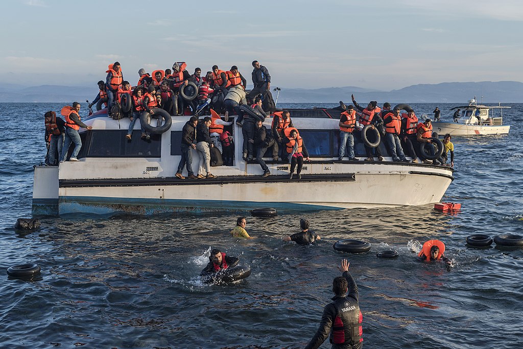 Vluchtelingen in reddingsvesten op het dek van een kleine motorboot in nood en in het water, met andere boten op de achtergrond.