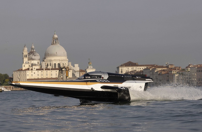 Un hydrofoil à moteur à l’allure futuriste sur l’eau avec la basilique Santa Maria della Salute de Venise en arrière-plan. 