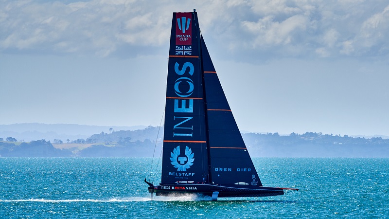 Modern AC75-class hydrofoil boat seen in profile with hull rising above bright blue sea, and hills in the background.