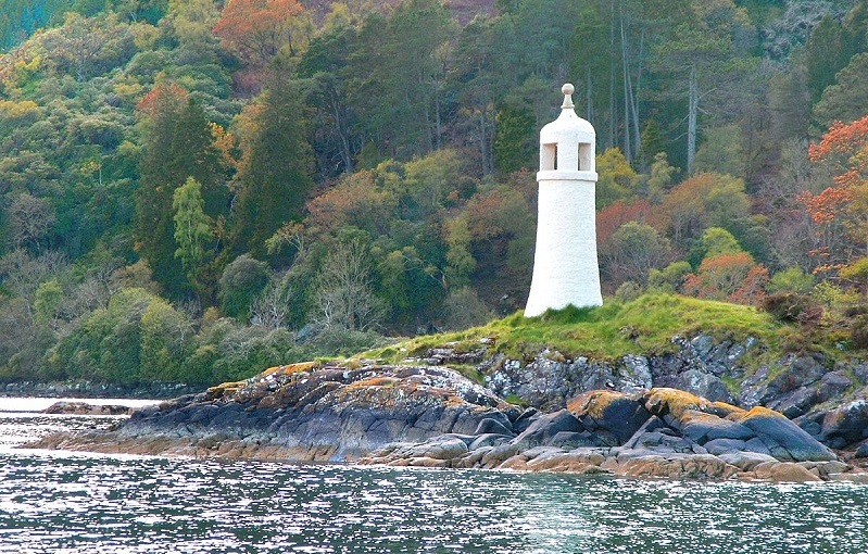 Ein kleiner, weißer Leuchtturm steht auf einem Felsen am Meer, umgeben von Wald.