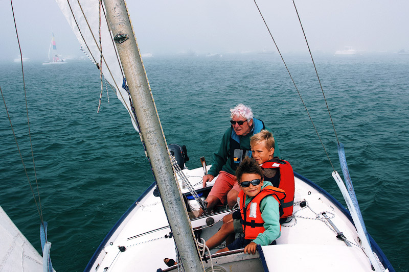 Un homme et deux jeunes enfants portant des gilets de sauvetage sont assis à l'avant d'un voilier.