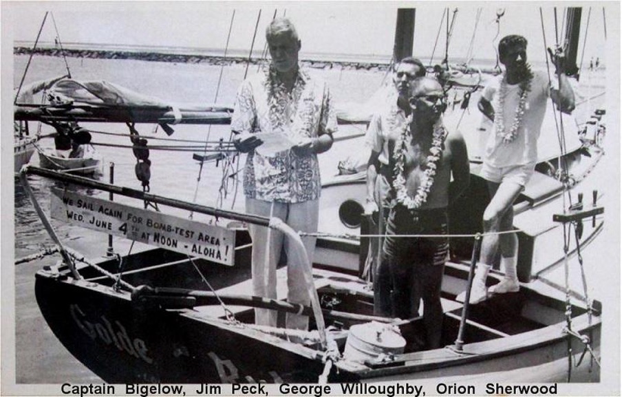 Oude zwart-witfoto van vier mensen aan boord van de Golden Rule. Aan de railing hangt een bord met de tekst ‘We sail again for bomb-test area…’.