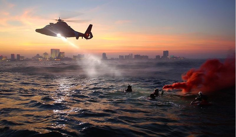 Helicopter shines light on group of people floating at sea with flare smoke, in front of sunrise over city skyline.