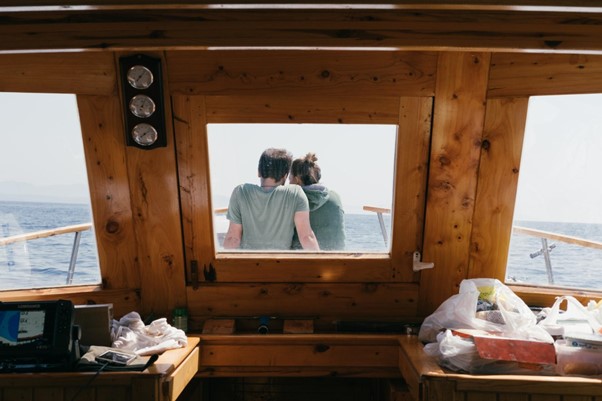 Man and woman sat outside on boat deck, seen from inside cabin with a lot of clutter visible.