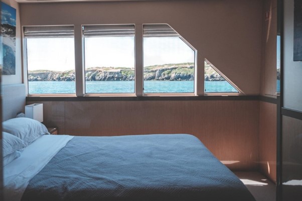 Neatly made bed in boat cabin with photograph on wall above headboard and coastline visible through window.