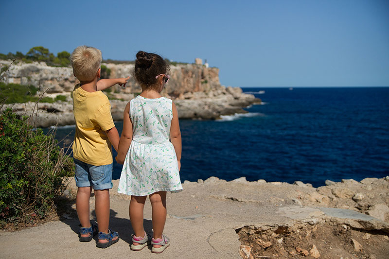Het achteraanzicht van een jongetje en een meisje die handjes vasthouden op de rand van een klif. Het jongetje wijst naar het uitzicht over de oceaan waar zich meer kliffen in de verte bevinden. 