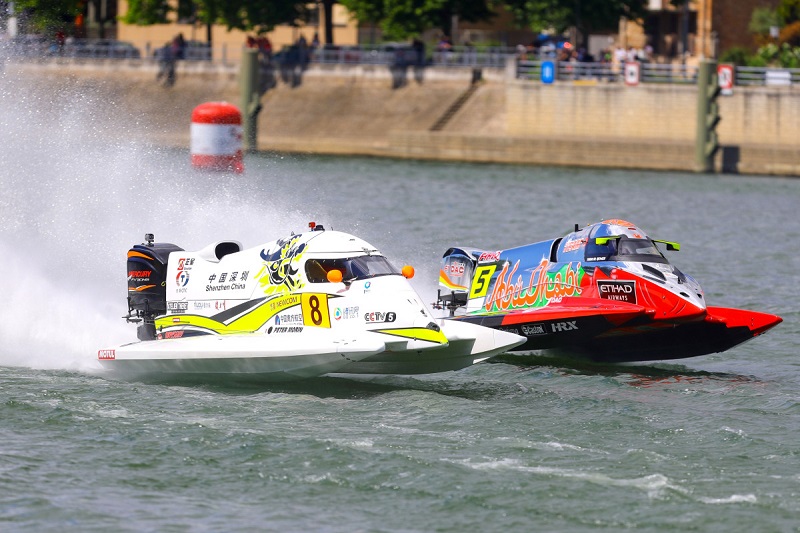 Two catamaran powerboats race alongside each other, with road in the background.