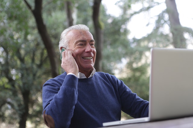 A man smiles at a laptop while holding his phone to his ear. 