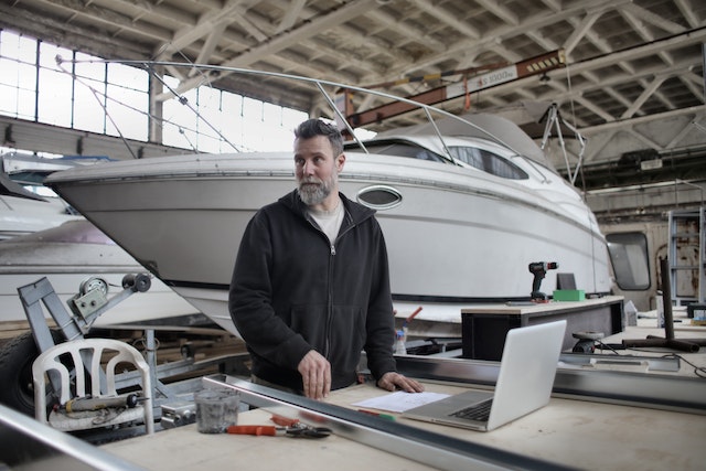 Man stands in front of laptop with several powerboats behind him inside a large warehouse.