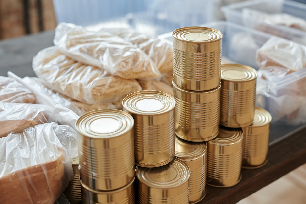 Una pila de latas sin abrir en primer plano y bolsas transparentes con pan al fondo 